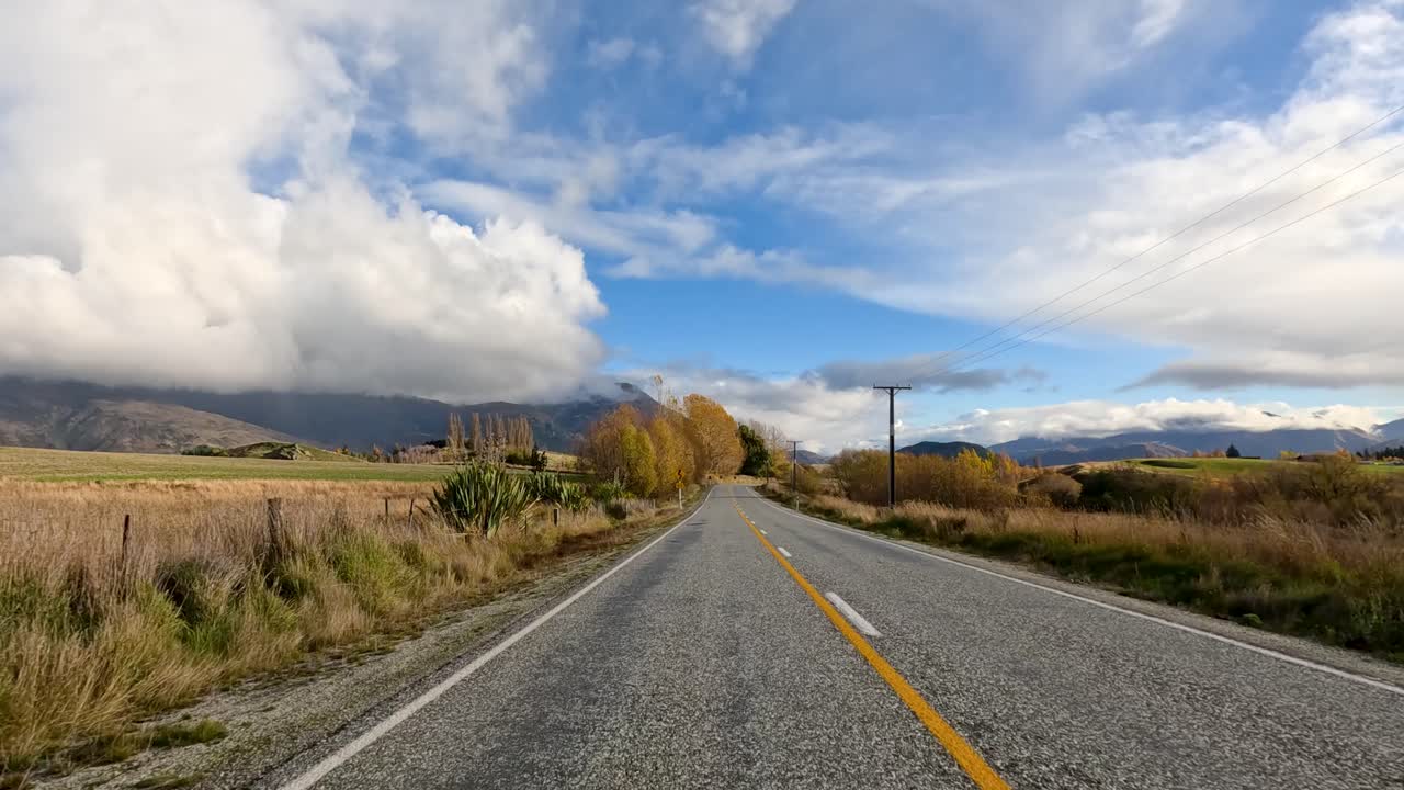 A vehicle travels along a quiet rural road bordered by fields, hills, and autumn trees under partly cloudy skies, with smooth forward camera movement