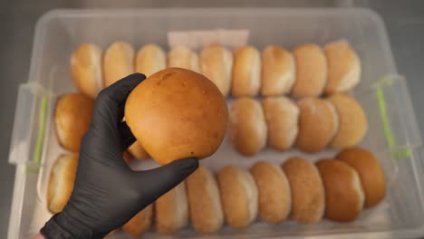 un recipiente de plástico con panes y una mano en un guante negro tomando un pan de él y sosteniéndolo en la palma