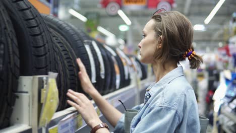 department of car accessories in the store. large hypermarket. the girl is standing near the rack with the tires. selects. blurred store background