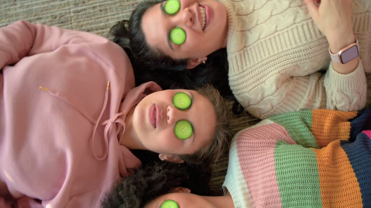 Diverse female friends lying on rug tilting, sharing glances under cucumbers while rotating camera