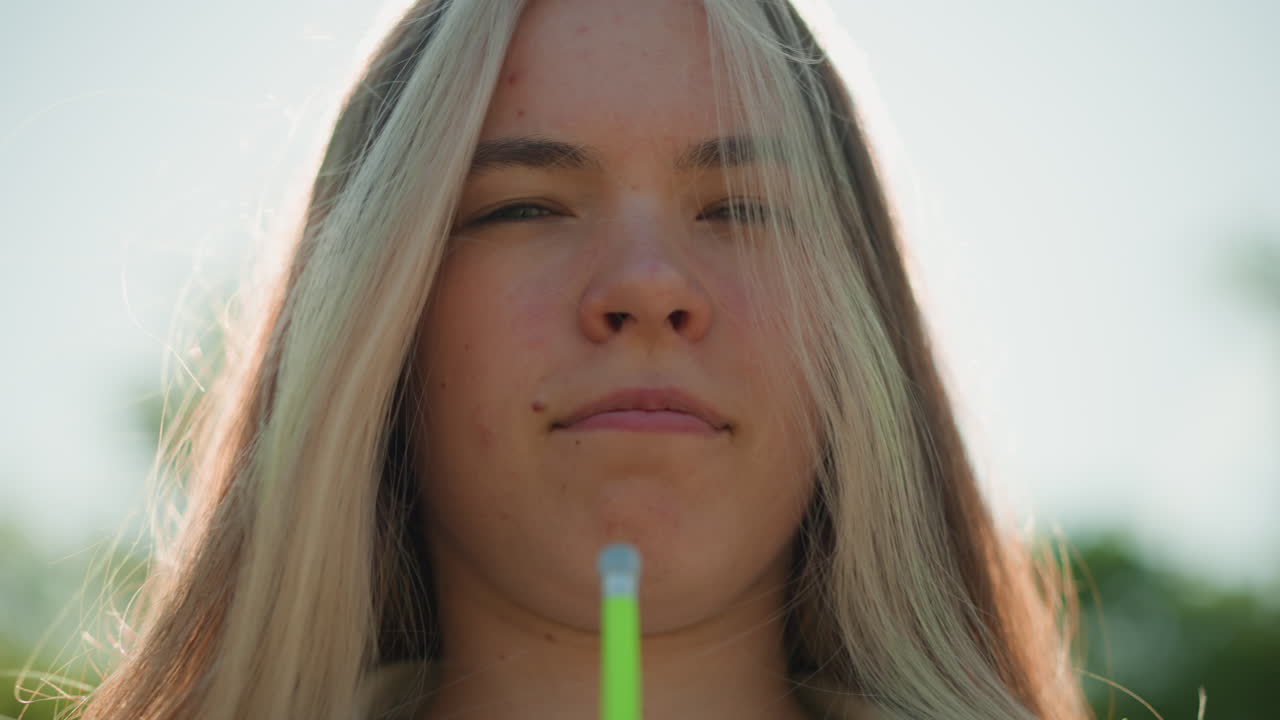 빛에 서 있는 젊은 여성의 클로즈업, 녹색 공기 프를 사용하여 그녀의 얼굴을 냉각하는 동안 그녀는 약간 눈을 감고, 그녀의 얼굴에 부드러운 바람을 느끼고, 빛이 필터링합니다.