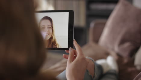 Woman-waving-hello-to-friend-on-vacation-at-home-through-internet-video-using-digital-tablet-app-on-sofa
