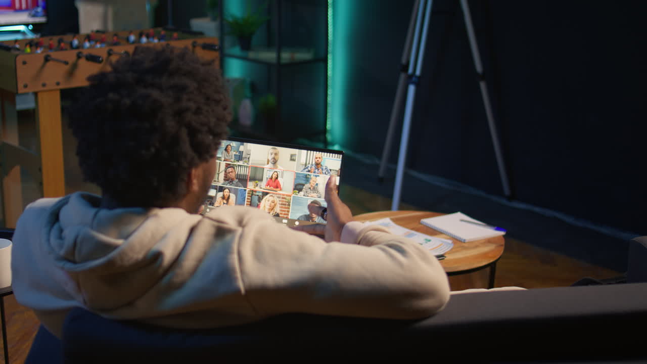 Focused young man smiling and waving during a web conference at his home, showcasing online communication in a digital and wireless lifestyle. Conference speaker with attendees. Camera B.