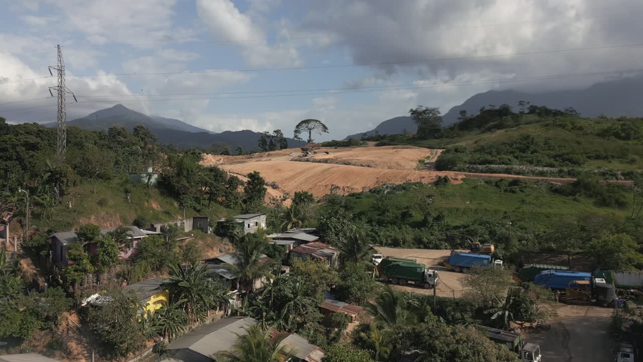 Wide shot of active excavation fields beside a rural neighborhood, with scattered houses, trucks, and distant mountain peaks under afternoon clouds