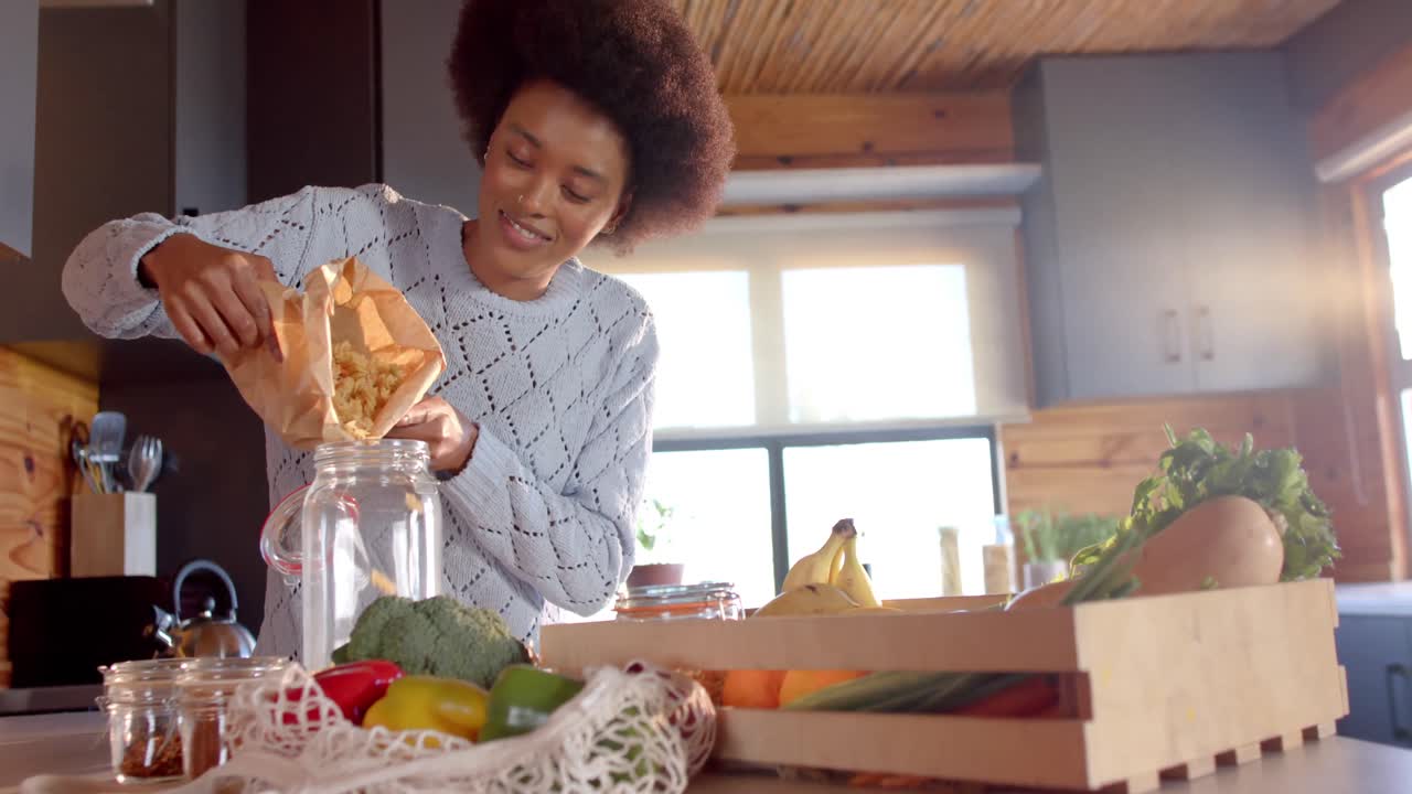 Happy african american woman pouring pasta into jar in sunny kitchen, in slow motion