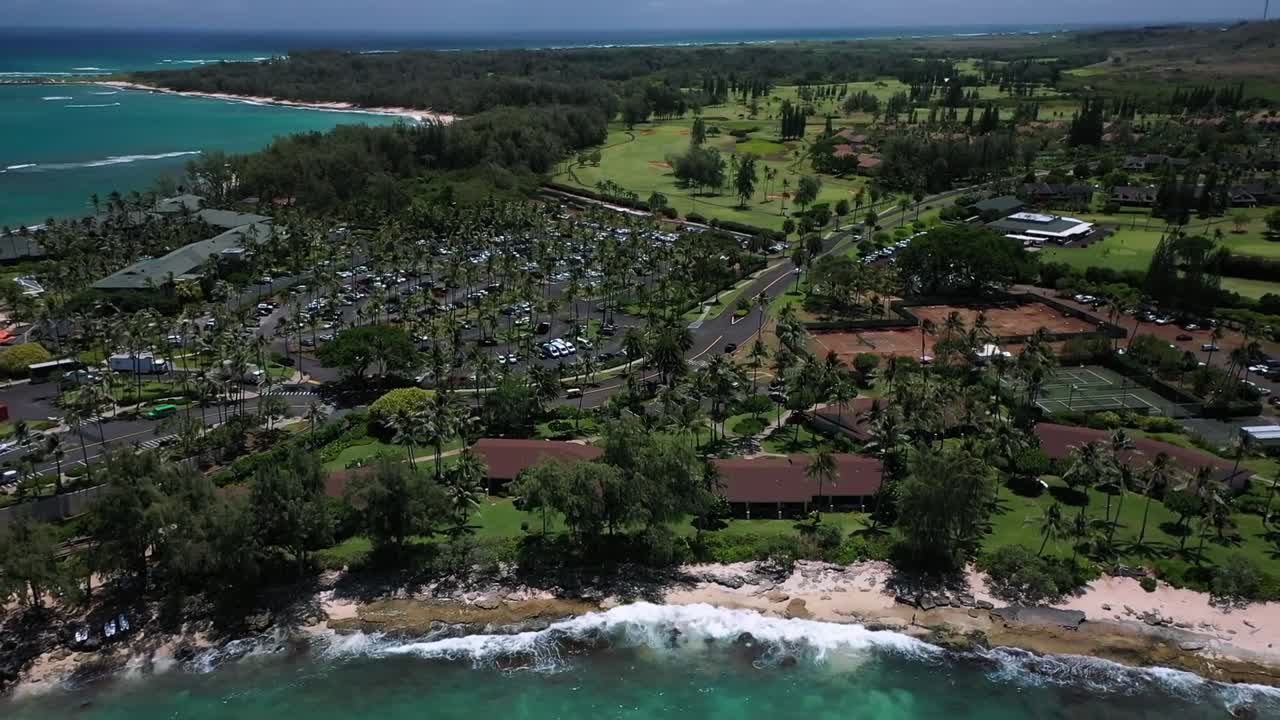 Aerial drone view of a tropical Hawaiian coastline featuring turquoise ocean waves, sandy beach, lush greenery, and resort-style buildings surrounded by palm trees under bright daylight