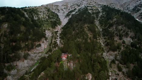 Un-Dron-Vuela-Alrededor-De-Una-Pequeña-Cabaña-De-Piedra-Rodeada-De-árboles-En-Un-Acantilado-Bajo-La-Cima-De-Una-Montaña