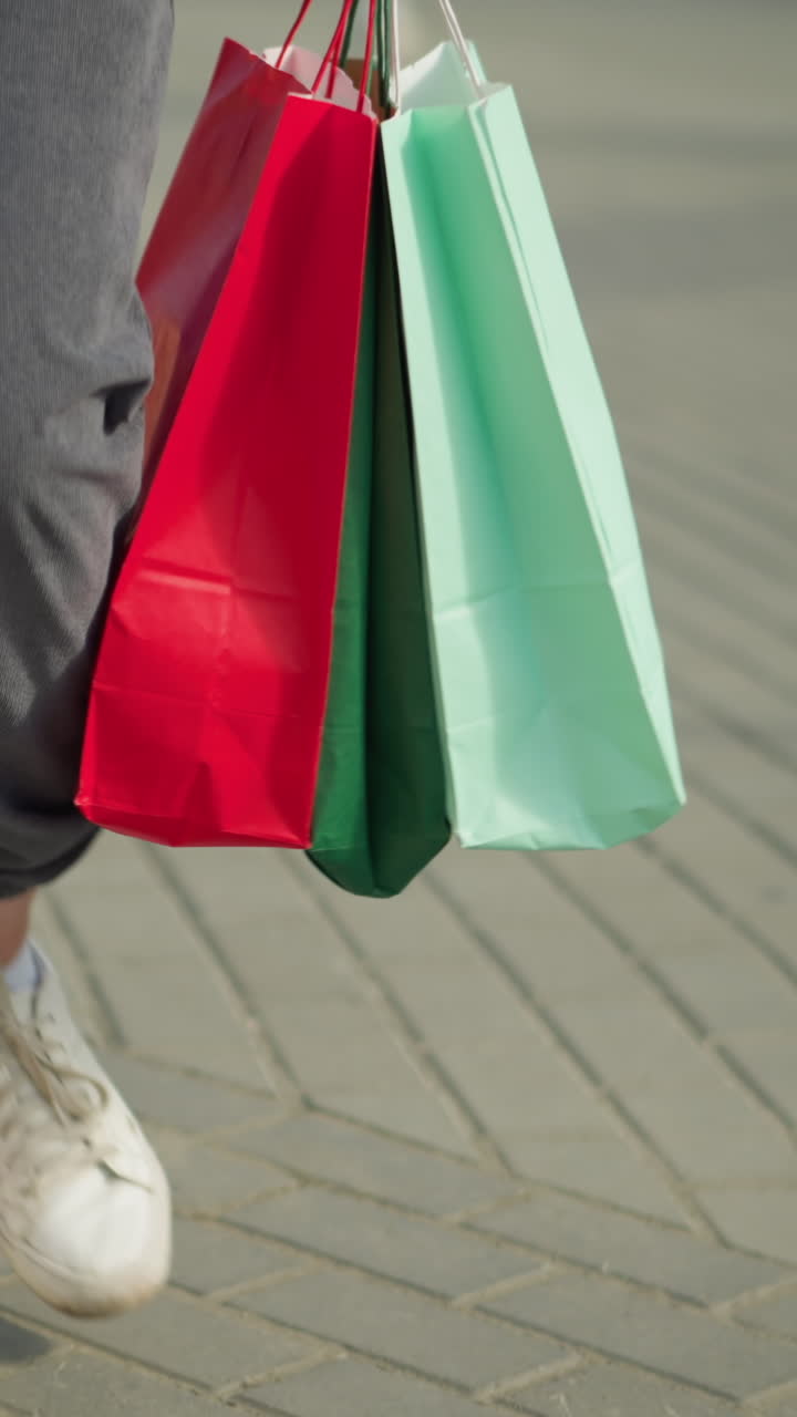 vista de la pierna de alguien con corredores grises y zapatos blancos de lona sosteniendo bolsas de compras verdes, menta y rojas mientras camina casualmente por un camino entrelazado, las bolsas se balancean con cada paso