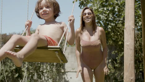 Feliz-Madre-En-Traje-De-Baño-Columpiándose-En-Un-Columpio-De-Madera-En-El-Patio-Trasero