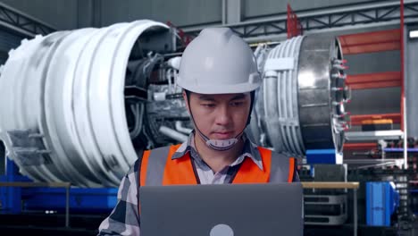 close up of asian male engineer with safety helmet working on a laptop while standing with airplane engine maintenance conducted, analytics checking the turbine
