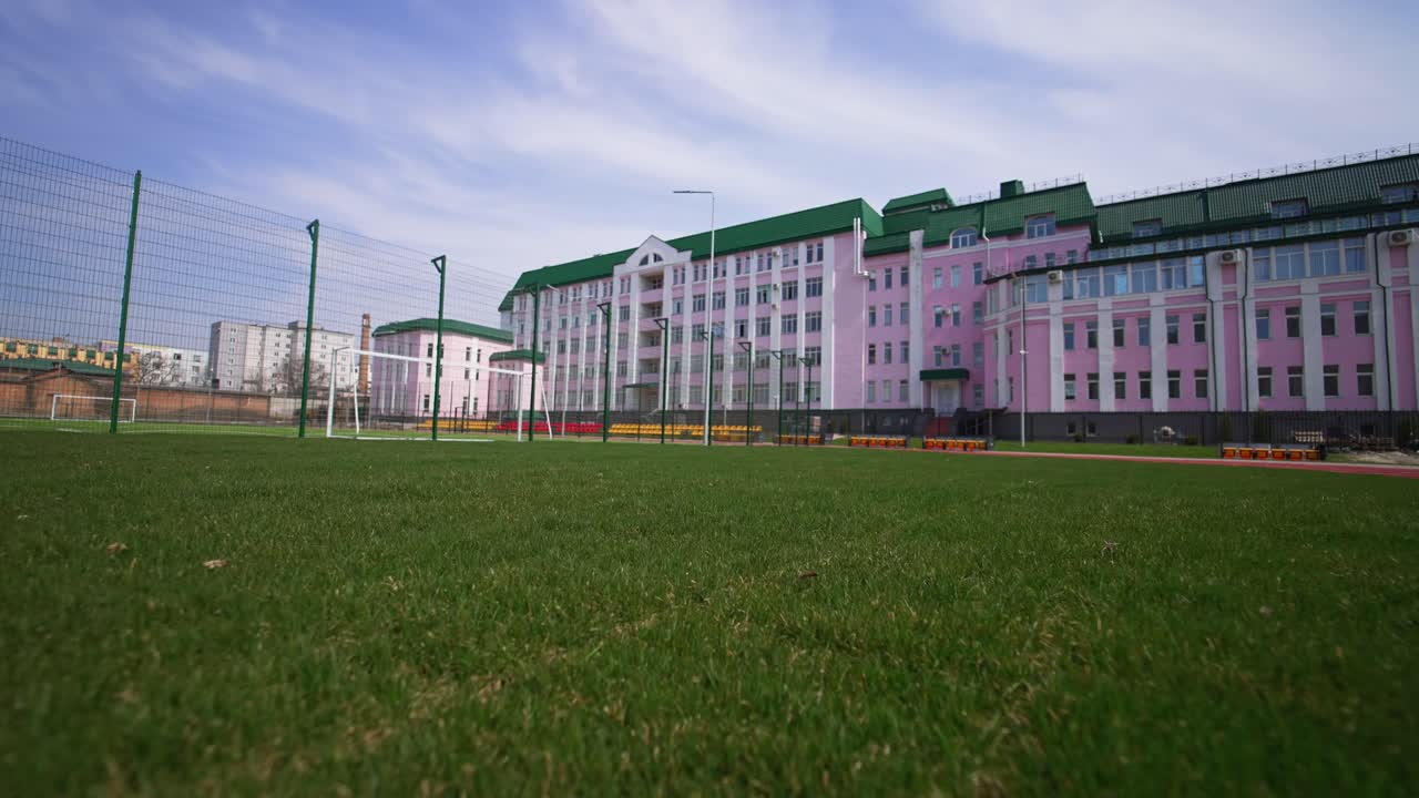 Red running track, green grass and football field located in front of the beautiful pink building. Low angle view. Blue sky at backdrop.