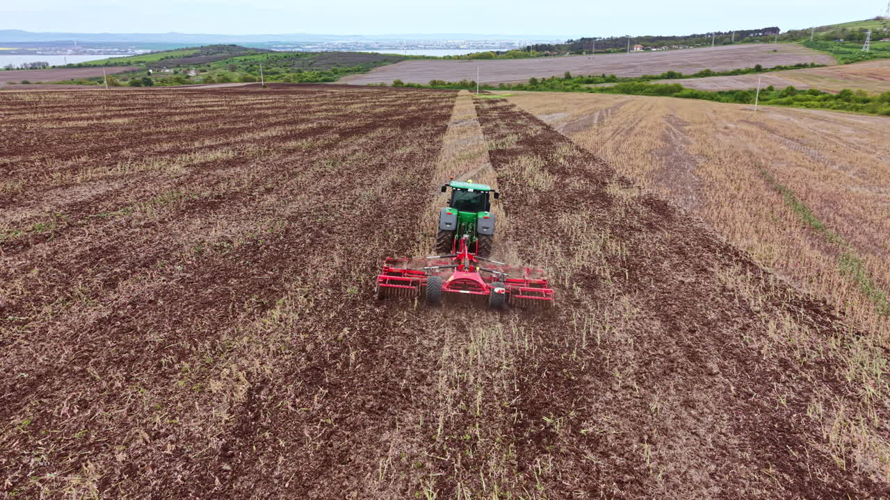Tractor preparando tierras de cultivo para la siembra durante el día en una zona rural cerca de colinas y una ciudad distante.