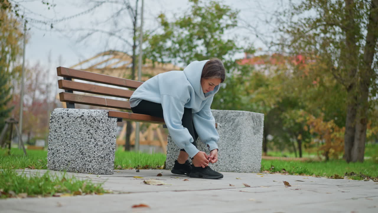 Girl sitting on concrete bench tightening shoelace in outdoor park, surrounded by trees and a pole in background, calm atmosphere, autumn setting, preparing for walk or exercise