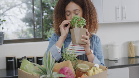 Happy-biracial-woman-unpacking-grocery-shopping-and-smelling-herbs-in-kitchen,-slow-motion