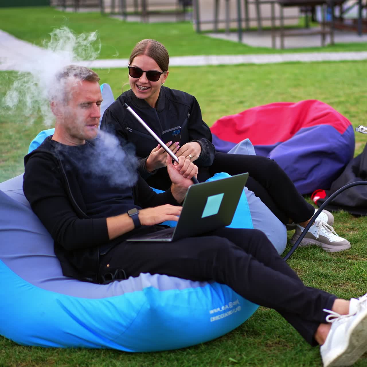 Young people in black clothes lie in bean bag chair relaxing outdoors. Man is working on laptop and smoking hookah and woman looks at her phone