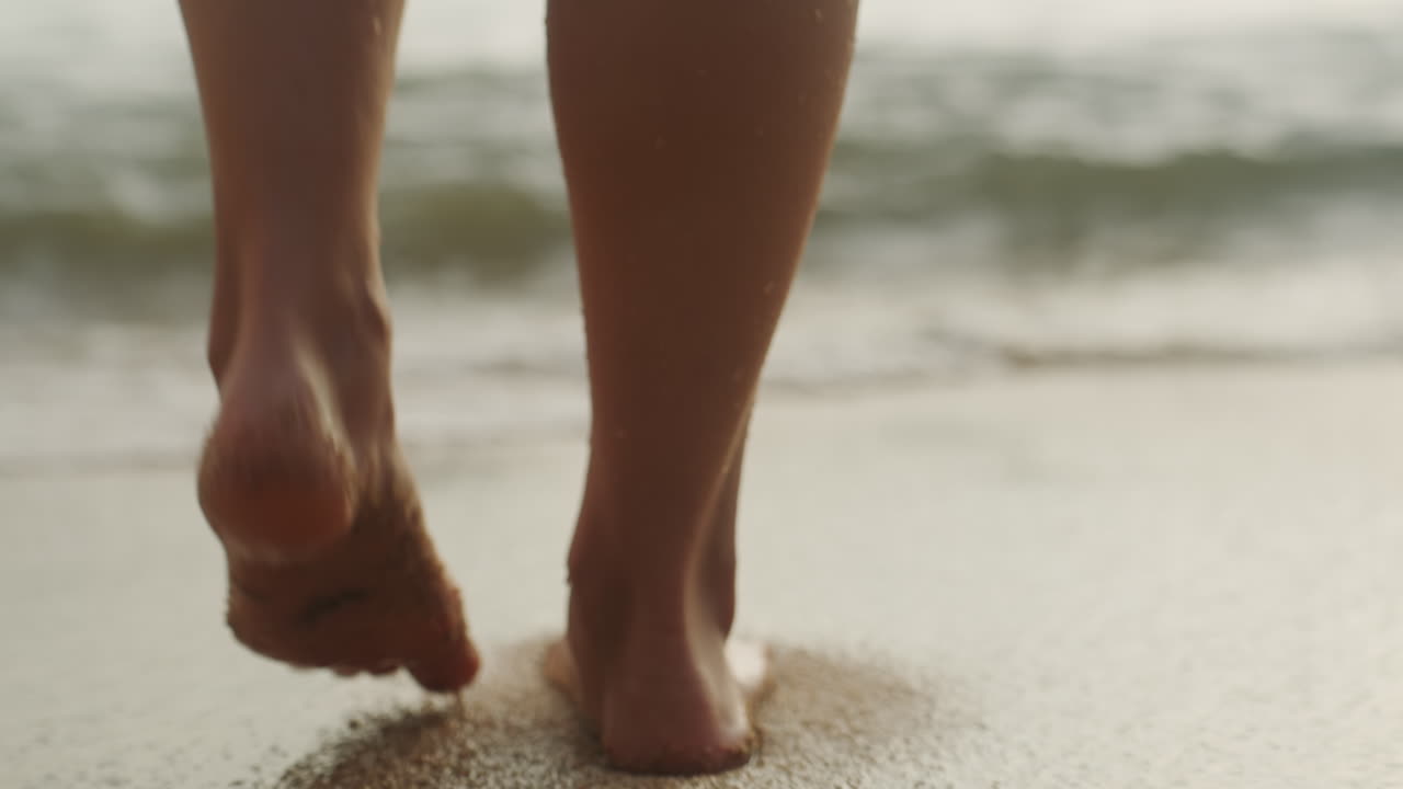 Passeggiata a piedi nudi lungo la riva sabbiosa, un viaggiatore mattutino che passa dalla sabbia asciutta alla risacca, goccioline sulla pelle, impronte leggere, ritmo giocoso, luce dorata dell'alba, movimento ravvicinato del piede.