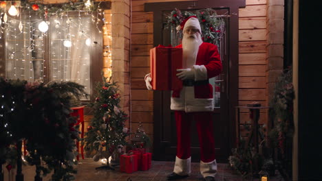 santa claus está de pie en el umbral de la casa con un regalo en sus manos en la víspera de navidad la cámara se acerca y santa pone su dedo a su boca y pide silencio