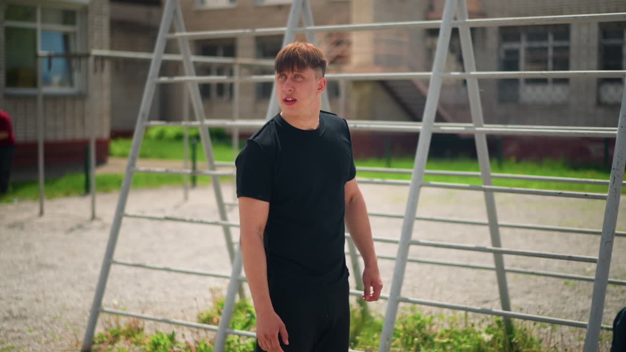 Hombre blanco joven con camiseta negra caminando cerca de unas barras metálicas, paseo pensativo por el campus con una bolsa informal, luz cálida del sol, patio residencial, postura atlética relajada, estudio sincero del movimiento