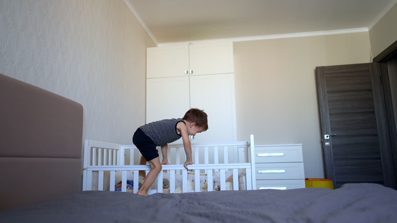 Toddler kid climbs from a big bed to his crib. Little baby boy hides in his bed. Active child playing at home.