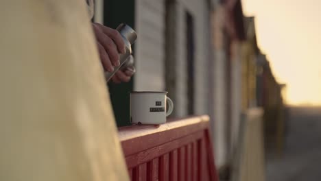 A-person-pouring-fresh-coffee-into-a-mug-while-in-a-beach-house-wooden-outdoor-deck