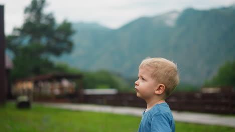 Upset-toddler-boy-stands-on-meadow-against-large-mountains