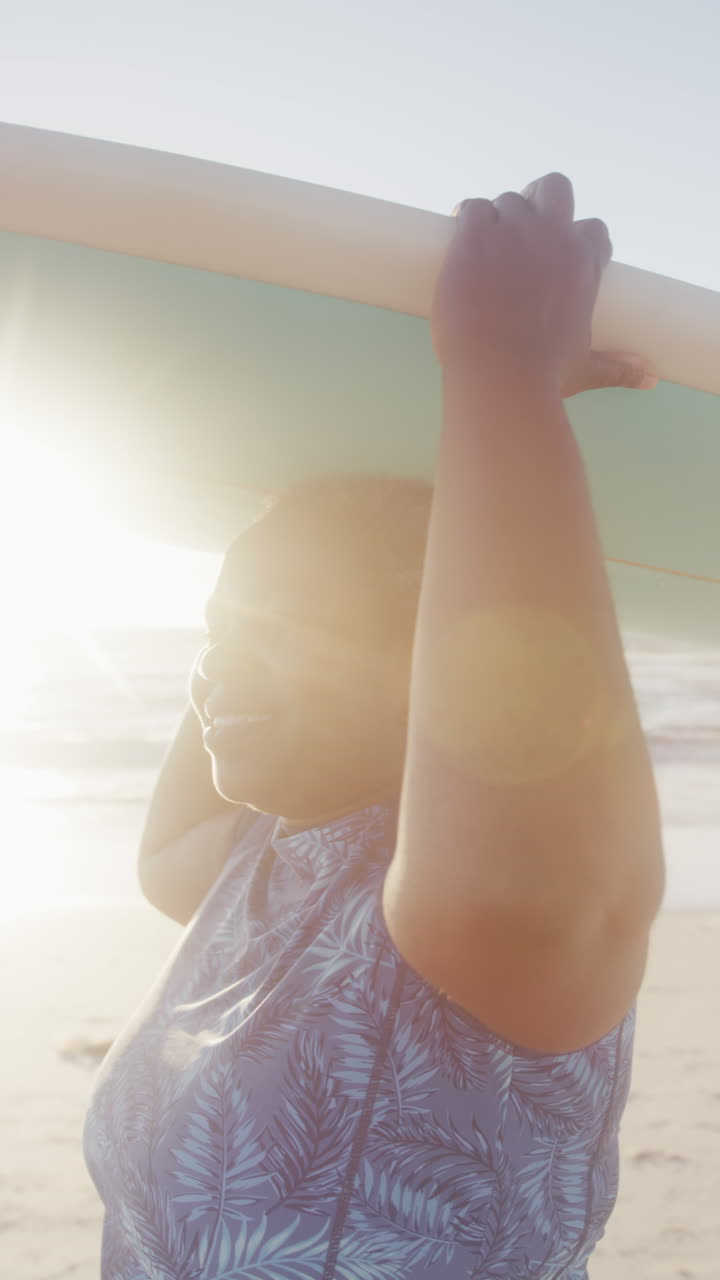 Vertical video of happy senior african american woman holding surfboard at beach, in slow motion