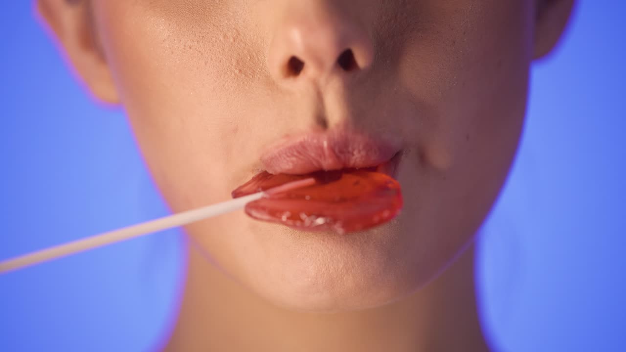 Medium shot of young woman lips licking a heart shaped lollipop while smiling sexy into the camera in slow motion against blue background