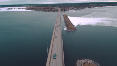 Toma-Aérea-De-4k-De-Autos-Que-Conducen-Sobre-Agua-Helada-En-Un-Enorme-Puente-De-Carretera-Rodeado-Por-Un-Gran-Lago-Congelado-De-Color-Verde-Azulado-Prores