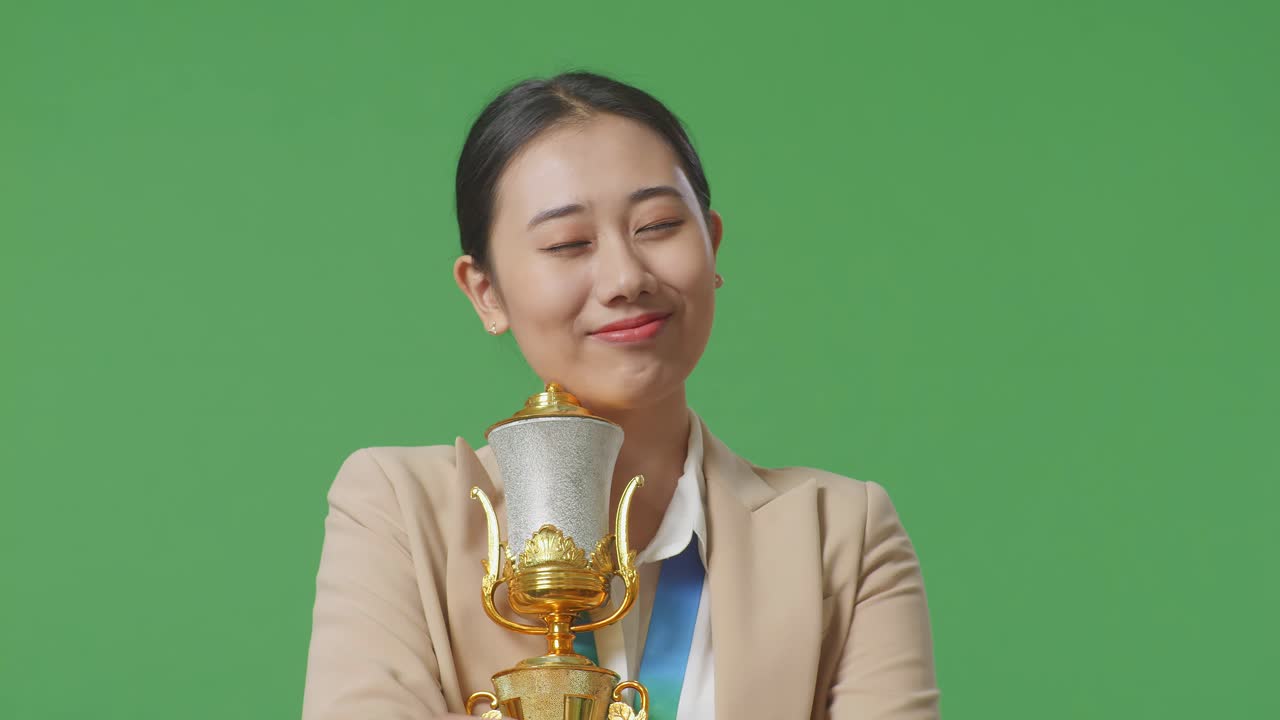 금메달을  슈트를 입은 아시아 사업가 여성이 황금 트로피를 안고 스튜디오의 녹색 스크린 배경에서 첫 번째 우승자로 자랑스럽게 승리하고 있습니다.