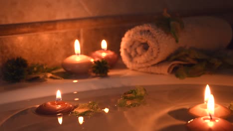 relaxing-spa-background-with-candles-floating-in-the-bath-water,-some-green-petals-and-a-towel-near-the-water-surface