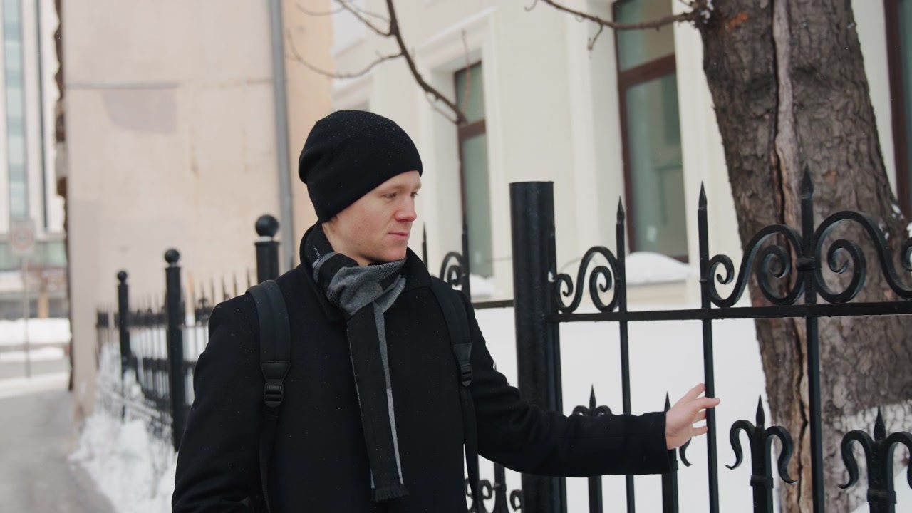 Close up of man wearing black coat and scarf touching wrought iron fence in snowy urban setting with white building background. Man remains focused on gate throughout emphasizing interaction