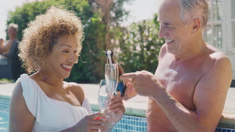 retired senior couple relaxing in swimming pool on summer vacation celebrating opening champagne