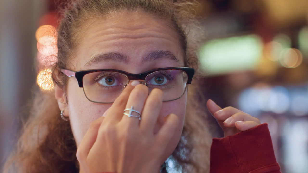 Jeune femme latine ajustant ses lunettes en intérieur. Gros plan montrant des expressions pensives, le bout du doigt touchant les montures, ajustement sur le nez, bague au doigt, lumières bokeh chaudes, intérieur d'arcade, étudiante.