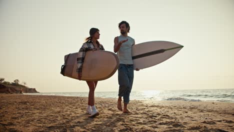 a brunette man with a beard in a white t-shirt walks with a blonde girl in a checkered shirt and tells her something, gesticulates with his hand, the couple carries surfboards and walks along the sandy seashore in the morning at sunrise