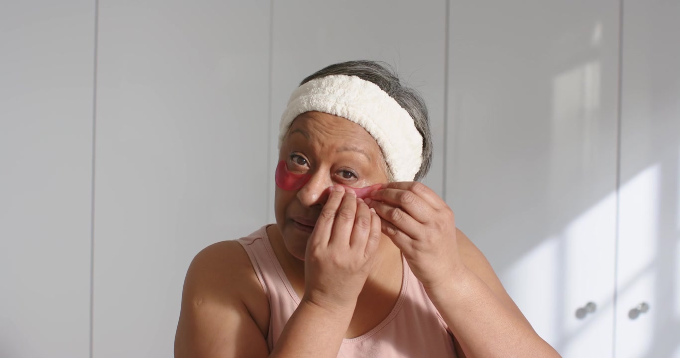 Iniciando a rotina de cuidados com a pele, mulher afro-americana idosa aplicando adesivos vermelhos para os olhos na penteadeira para reduzir bolsas