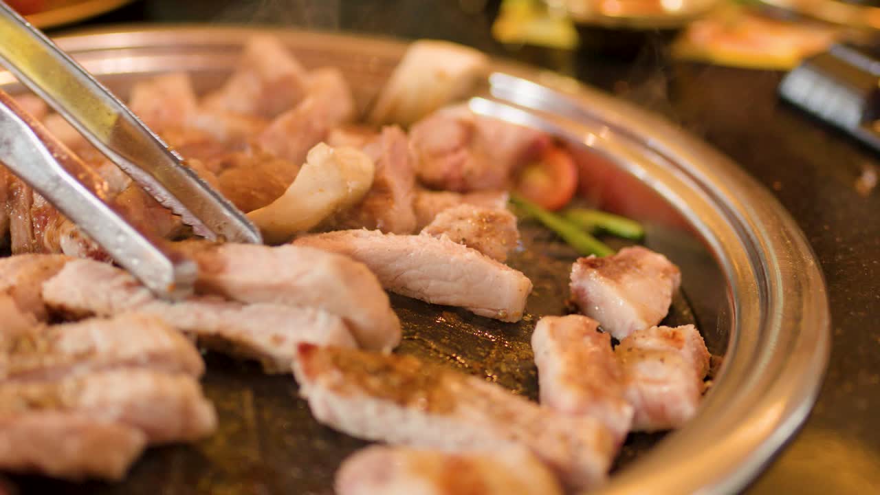 Juicy pork belly slices grilled on round metal pan, tongs turning meat, warm indoor lighting