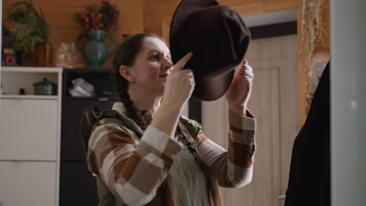 Window view shows woman approaching corner where jacket and hat hang on hook then picking hat and wearing it on head in natural light inside wooden room while dressed in plaid shirt and olive gown
