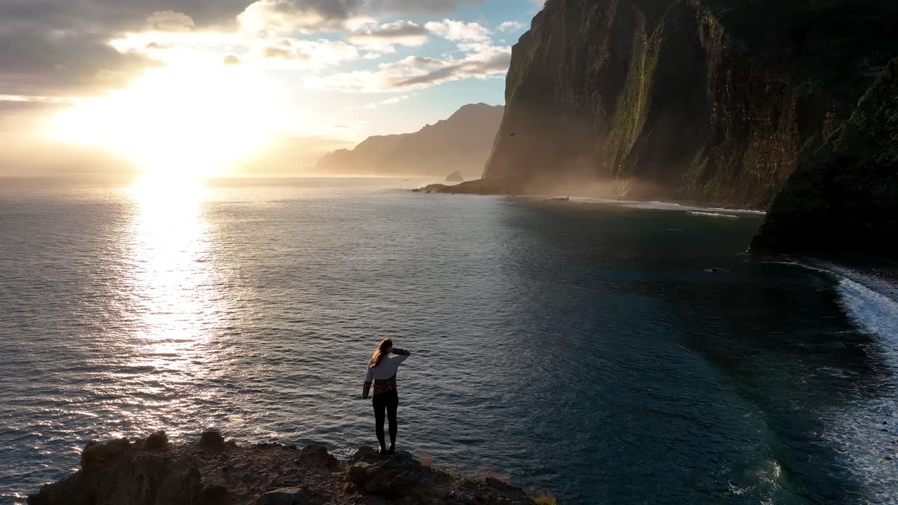 antena de una mujer de pie en lo alto de un acantilado rocoso con vistas a la puesta de sol costera y el sobrevuelo de aves, madiera, portugal