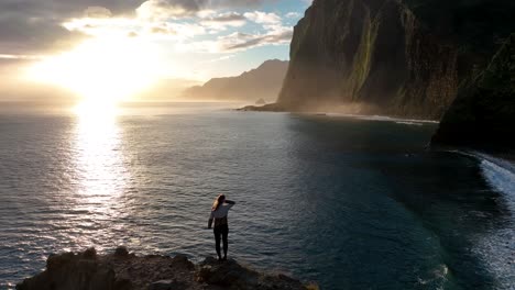 Aerial-of-woman-standing-ontop-of-rock-cliff-overlooking-coastal-sunset-and-bird-flyby,-Madiera,-Portugal