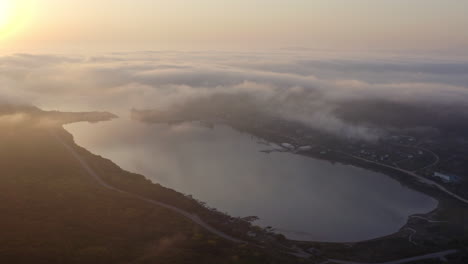 Volar-Sobre-Una-Bahía-Con-Agua-Tranquila,-El-Sol-Reflejándose-En-La-Superficie-Del-Agua-Y-Una-Densa-Nube-De-Niebla-Proveniente-De-Las-Colinas,-Al-Atardecer