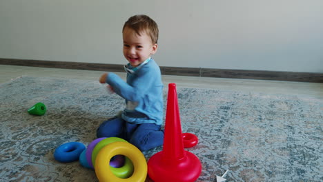 energetic caucasian toddler collecting toy pyramid. smiling kid turns and waves his hands scaring the black cat.