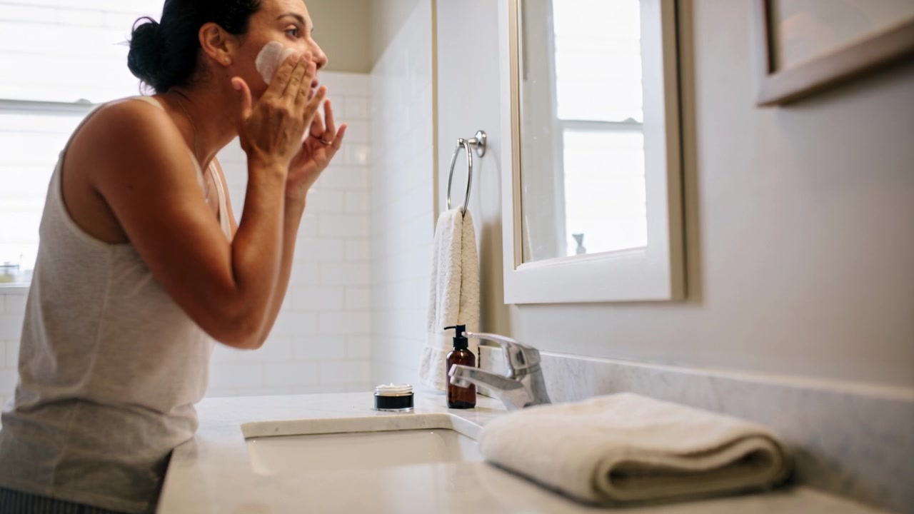 Leaning woman in tank top looking into mirror, applying cleanser at sink for facial cleansing