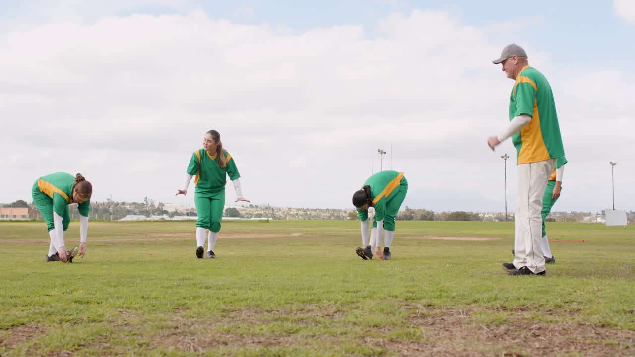 Multiracial female baseball players and male coach warming up and stretching legs on a pitch