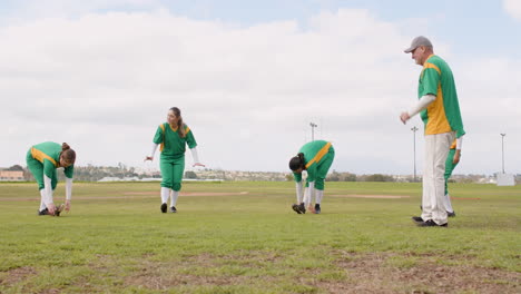 multiracial female baseball players and male coach warming up and stretching legs on a pitch