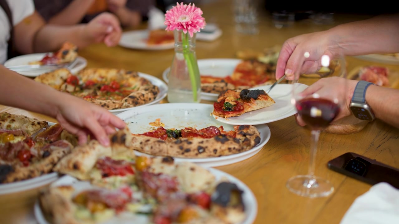 Primo piano di persone che condividono pizza artigianale e vino rosso su un tavolo di legno.