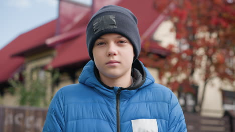 boy wearing black beanie and blue jacket standing outdoors, looking thoughtfully in front of beautiful autumn trees with colorful red leaves and a house in background