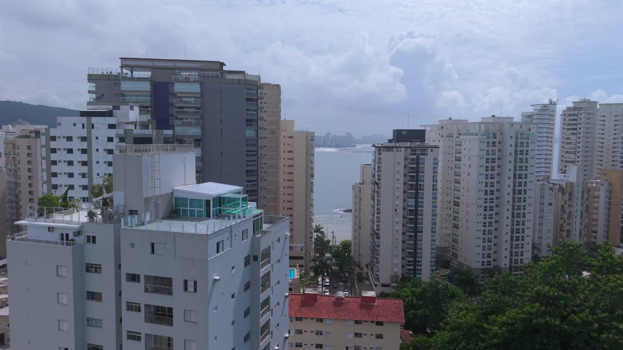 Praia das Astúrias no Guarujá, São Paulo, Brasil, revelada através da Mata Atlântica costeira com edifícios altos densos e torres com vista para o mar, paisagem costeira urbana, aproximação de drone