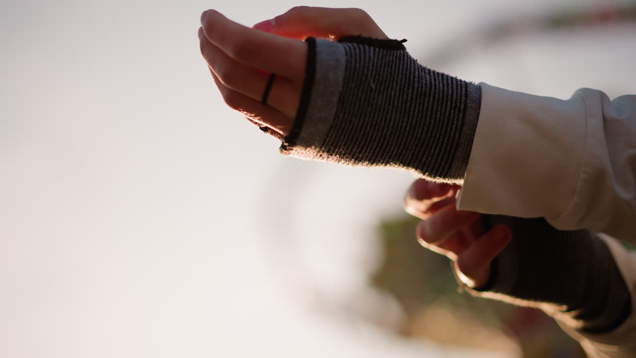 Nahaufnahme von Händen, die bei Sonnenuntergang einen Handschuh anziehen, hintergrundbeleuchtetes Riesenrad und warmes goldenes Licht, Gitarrist, der fingerlose Strickstulpen mit beringten Fingern über das Handgelenk zieht, taktile Stoffdetails, ruhige Probe