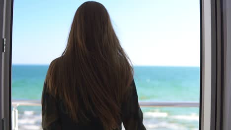 Woman-with-long-hair-opening-balcony-doors-and-looking-out-at-the-sea.-Enjoying-the-sea-view-outside.-Slowmotion-shot