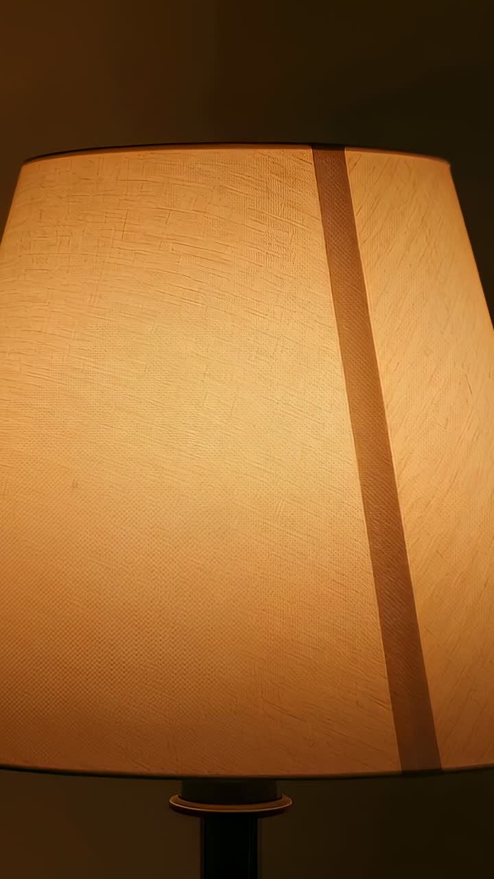 Vertical video: Shifting camera framing nudging lit table lamp shade at home, showing beige seam
